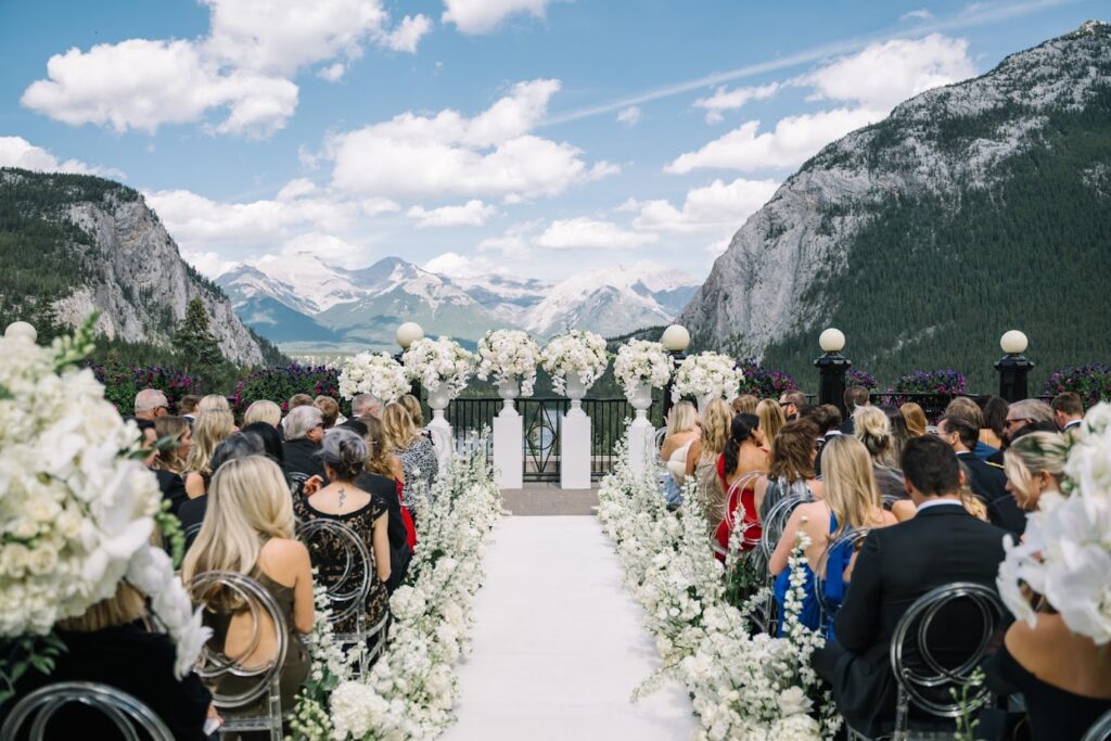 Outdoor wedding ceremony at Fairmont Banff Springs, overlooking the lake and the Rocky Mountains in the background, the aisle is lined with all white flowers
